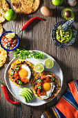 Huevos Rancheros are Mexican Ranchero Eggs breakfast with pico de gallo and guacamole sauces, hot chili pepper, tortillas on rustic wood