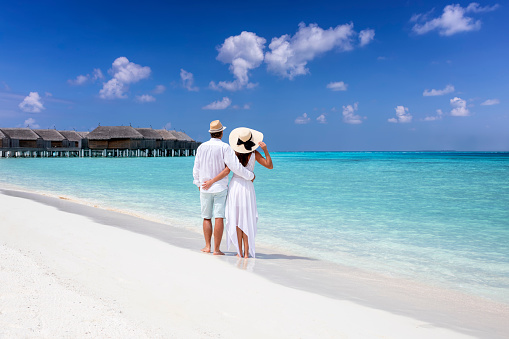 A hugging couple in white summer clothing stands on a tropical beach and enjoys the view to the turquoise sea and blue sky