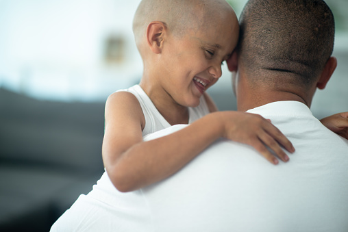 A father is seen from behind with his son wrapping his arm around him to pull him in for a hug.  He is dressed casually in a white tank top and smiling at his father.