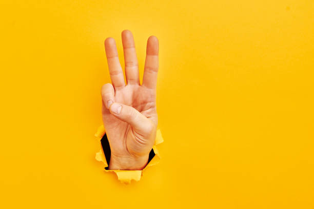 Human hand reaching through torn yellow paper sheet showing number three stock photo