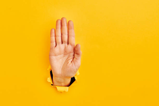 Human hand reaching through torn yellow paper sheet showing "stop sign" stock photo
