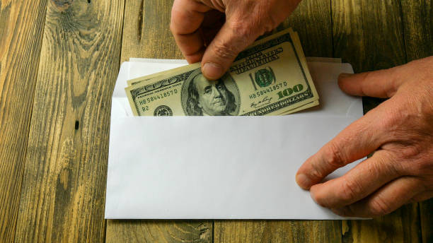 Human hands put two one hundred dollar bills in cash into white paper envelope that lies on brown wooden table. Concept of saving money or corruption and shadow economy. Human hands put two one hundred dollar bills in cash into white paper envelope that lies on brown wooden table. Concept of saving money or corruption and shadow economy. Close-up. economic stimulus stock pictures, royalty-free photos & images