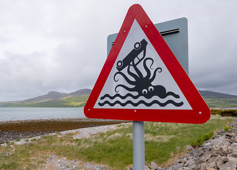 A humorous road sign beside a causeway near the coastal village of Tongue in the northwest of Scotland.  The warning sign notifies drivers that they are going to be driving close to the sea. The black and white illustration is of an octopus holding an upturned car in its tentacles.