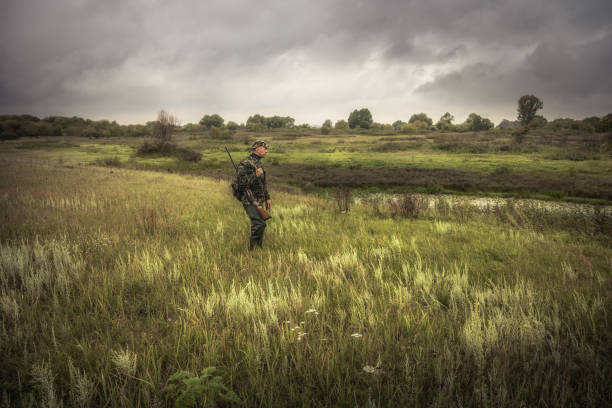 Hunting hunter men with gun standing in field nearby river during hunting season in overcast day stock photo