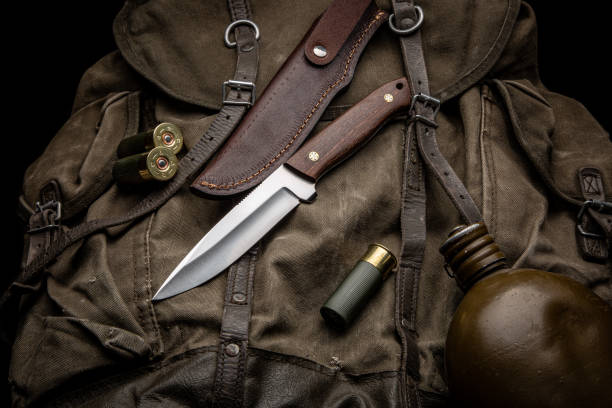 Hunting knife with a wooden handle on a vintage canvas backpack. 12-caliber shotgun shells and a water canteen are nearby. Hunter's set. Dark background. stock photo