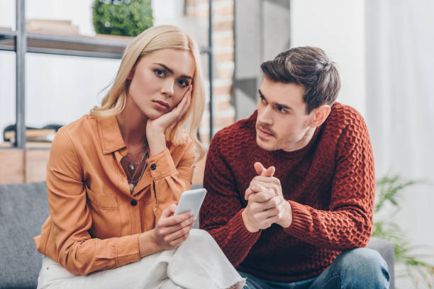 husband looking at unhappy young woman holding smartphone and looking at camera at home, relationship problem concept husband looking at unhappy young woman holding smartphone and looking at camera at home, relationship problem concept envy stock pictures, royalty-free photos & images