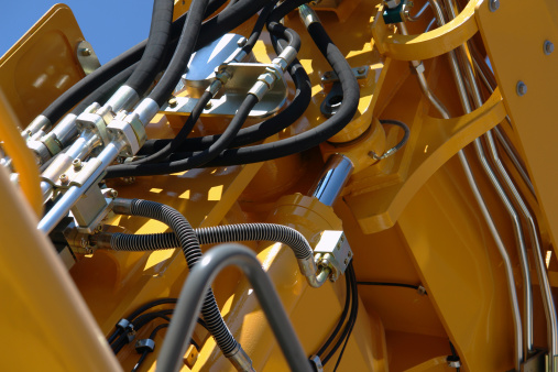 This is a technical closeup from the hydraulic system of a new, big excavator. There are many black hoses visible in the photo, and the yellow coated metal reflects the sun. The geometry visible in the photo's composition is quite complex.
