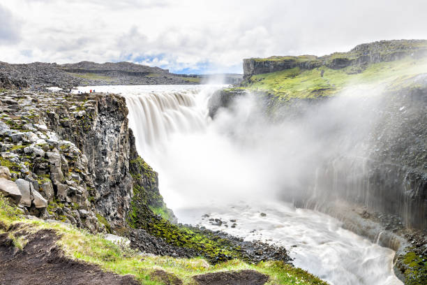 Icelandic Dettifoss waterfall, Iceland, largest volume in Europe, gray grey water, rocky cliff, rocks, soil, green grass, water flowing mist spraying, people walking  dettifoss waterfall stock pictures, royalty-free photos & images