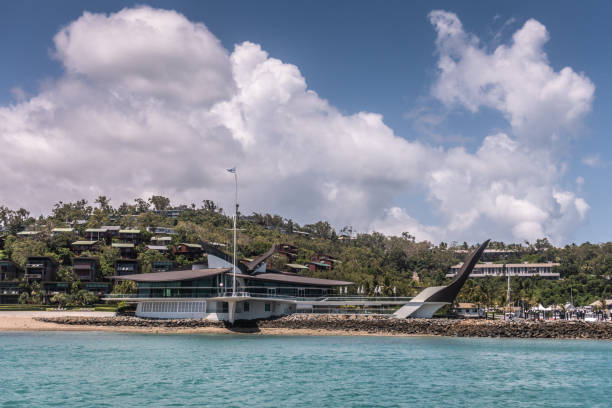 Iconic Yacht Club House shot from sea in marina , Hamilton Island, Australia. Hamilton Island, Australia - February 16, 2019: Wider shot of Black and white Iconic Yacht Club House, shot from sea, under blue sky and white cloudscape. Green hill with housing in back. unesco organised group stock pictures, royalty-free photos & images