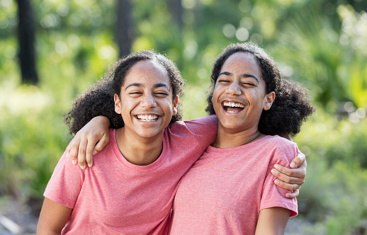 Identical twin teenage girls wearing pink t-shirts, standing side by side outdoors, grinning with their arms around each other's shoulders. The African-American sisters are 14 years old.