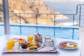 Horizontal color close-up image of delicious breakfast arranged on table. Private swimming pool and sea in the background.