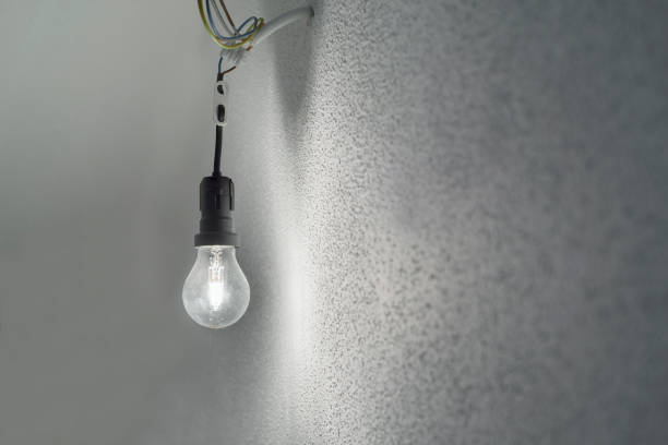 Illuminated halogen incandescent light bulb in a black plastic lamp holder socket with power cables protruding from a wall with white rough plaster stock photo