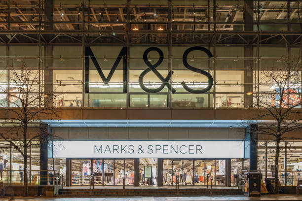 Illuminated night view of British retailer M&S Group plc closed shop entrance with logo & sign at the city center. stock photo