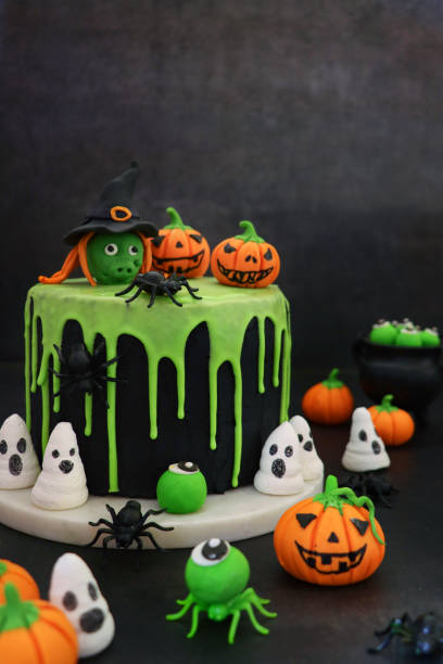 Image homemade, Halloween, layered sponge cake on marble cake stand, covered in black fondant icing with dripping green glace icing design, topped with edible pumpkins and witch, eyeball, ghost meringue kisses and plastic spiders, black cauldron stock photo