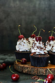 istock Image of batch of homemade, Black Forest gateau cupcakes in brown paper cake cases on wooden cake stand, piped whipped cream rosettes topped with morello cherries sprinkled with chocolate shavings, black background, focus on foreground 1326614660