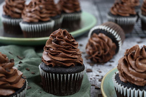 Stock photo showing a close-up view of freshly baked, homemade, chocolate cupcakes in paper cake cases on a green plate. The cup cakes have been decorated with a swirl of chocolate piped icing and topped with chocolate pieces.
