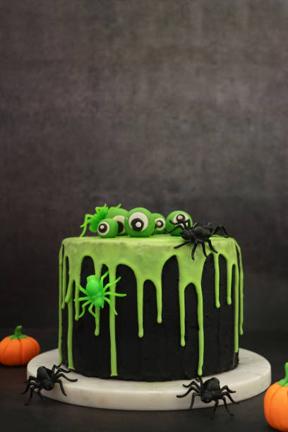 Image of homemade, Halloween, layered sponge cake on marble cake stand beside fondant pumpkins, covered in black fondant icing with dripping green glace icing design, topped with edible eyeballs and plastic spiders, black background, focus on foreground stock photo