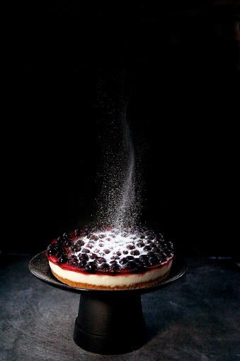 Stock photo showing close-up view of icing sugar onto blackcurrant cheesecake with buttery biscuit base, vanilla mascarpone cream cheese for dessert pudding on a black cake stand.
