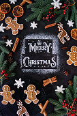 istock Image of Merry Christmas message stencilled, white icing sugar on black background, surrounded by reindeer and gingerbread men cookies, fondant icing snowflakes, dried citrus orange fruit slices, star anise, cinnamon sticks, spruce needles and red berries 1355500618