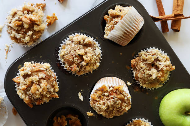 Image of muffin baking tray with rows of homemade apple muffins in paper cake cases topped with streusel crumble on marble effect background, elevated view stock photo