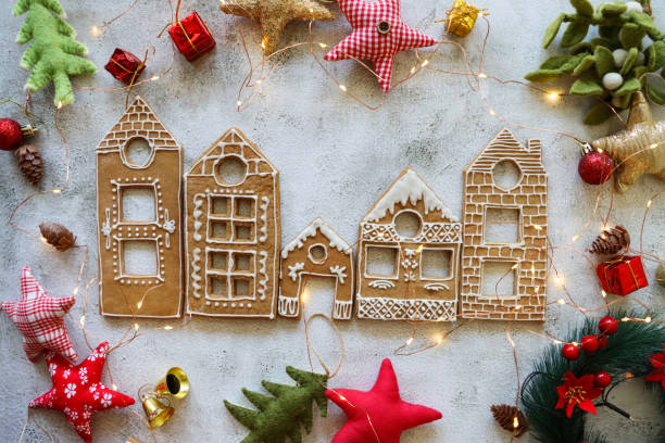 Image of row of homemade, house-shaped, gingerbread biscuits, cookies iced with white glace icing, Christmas village display surrounded by felt Christmas tree and star-shaped ornaments, marble effect background, elevated view stock photo