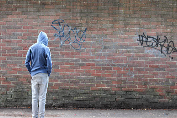 Image of teenage boy / youth wearing hoodie, beside graffiti wall Photo showing a teenage boy / youth wearing a blue hoodie and standing beside a brick wall, in a run-down part of the city. vandalism stock pictures, royalty-free photos & images