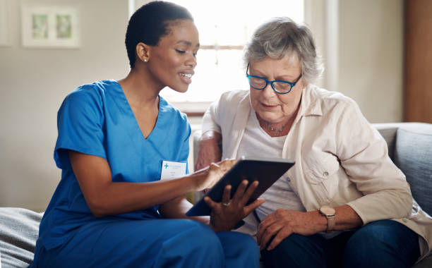 Improving the quality of healthcare through with digital tech Shot of a senior woman using a digital tablet with a nurse on the sofa at home home caregiver stock pictures, royalty-free photos & images