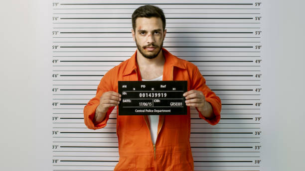 In a Police Station Arrested Man Getting Front-View Mug Shot. He's Wearing Prisoner Orange Jumpsuit and Holds Placard. Height Chart in the Background. In a Police Station Arrested Man Getting Front-View Mug Shot. He's Wearing Prisoner Orange Jumpsuit and Holds Placard. Height Chart in the Background. police force photos stock pictures, royalty-free photos & images