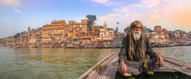 Indian sadhu (monk) enjoy boat ride at Varanasi Ganges river with view of ancient Varanasi city architecture and ghat Sadhu baba sitting on a wooden boat overlooking ancient Varanasi city architecture with Ganges river ghat at sunset. ghat stock pictures, royalty-free photos & images