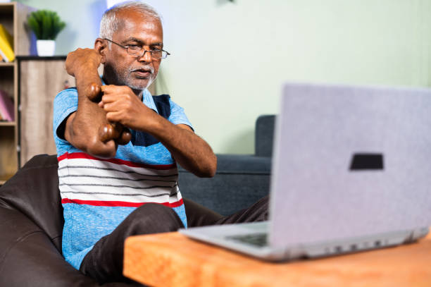 indian senior old man massaging hand using wood roller massager by watching online medication videos on laptop from home - concept of physiotherapy, technology and healthy lifestyle. indian senior old man massaging hand using wood roller massager by watching online medication videos on laptop from home - concept of physiotherapy, technology and healthy lifestyle doctor of physio therapy online stock pictures, royalty-free photos & images
