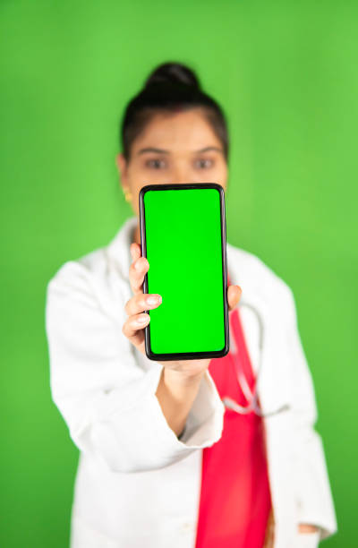 Indian young female doctor holding smartphone against green background. Happy Indian young female doctor holding touch screen smartphone and standing against an isolated green background. smart phone green background stock pictures, royalty-free photos & images
