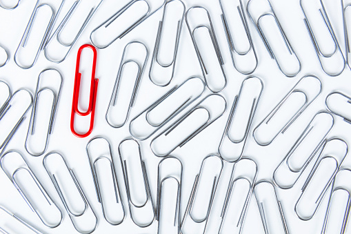 Abundance of silver colored paperclips where only one is red on white background.