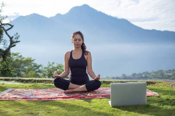Indonesian Woman Meditating in Nature Close-up shot of Indonesian yoga instructor meditating in nature. She's doing live broadcast via laptop, sitting crossed legged above a rug on a grass , with a mountain background behind her. She's wearing a black sports clothing with a ponytail hair style. personal trainer online stock pictures, royalty-free photos & images