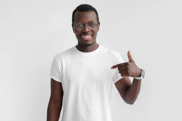 Indoor photo of young African American man pictured isolated on grey background pointing to his white blank T-shirt drawing attention to advertisement on it, promoting goods, apps or services Indoor photo of young African American man pictured isolated on grey background pointing to his white blank T-shirt drawing attention to advertisement on it, promoting goods, apps or services blank t shirt stock pictures, royalty-free photos & images