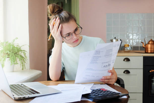Indoor shot of young European Caucasian girl looking at financia Indoor shot of young European Caucasian girl looking at financial documents at home with deeply bored face looking sick and tired of her economic problems, trying to check counts and all details debt stock pictures, royalty-free photos & images