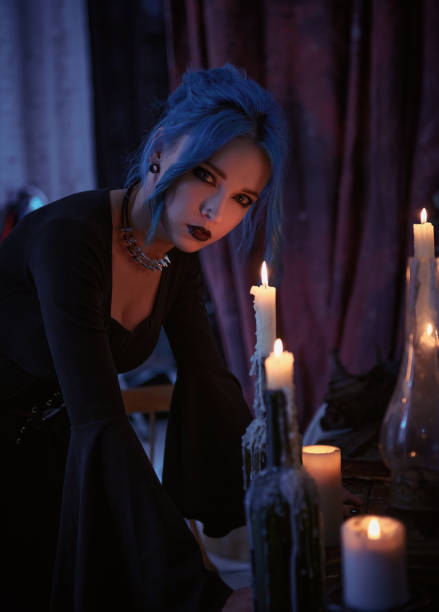 Indoors portrait of gorgeous goth girl in black dress. Blue-haired gothic lady among candles. Portrait of mysterious young witch stock photo