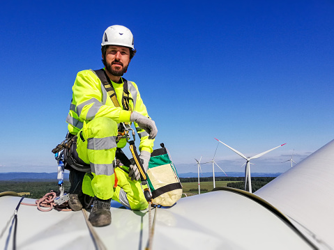 Industrial climber,  rope access technician kneeling on enormous onshore wind-turbine while preparing for blade inspection with wind farm behind him and looking into the camera