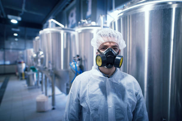 Industrial plant worker wearing mask and hazard suit for protection. Portrait of industrial worker technologist wearing hazmat suit in production plant. Man in white protective uniform with hairnet and protective mask handling hazardous chemicals. radioactive contamination stock pictures, royalty-free photos & images