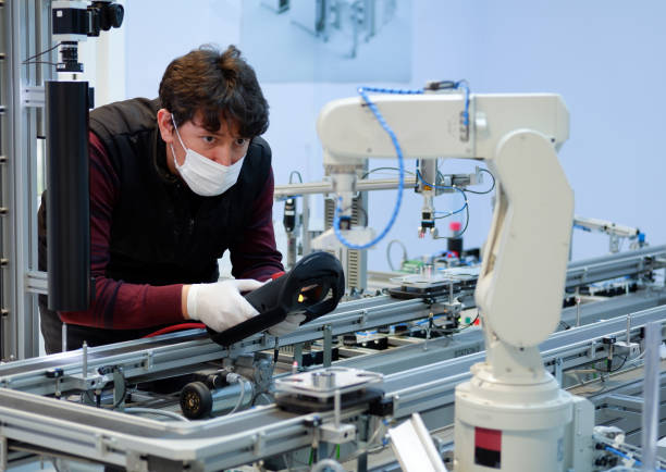 industry 4.0 : Man with surgical mask and gloves against COVID-19 ( Coronavirus ) programming robot arm with teach pendant. stock photo