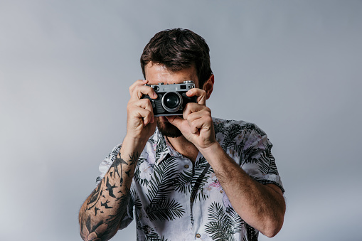Portrait of hipster young man wearing a Hawaiian shirt, holding a camera in hand, looking at the camera. Studio shot, one person, gray background.