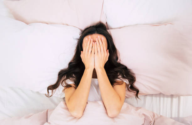 Insomnia! It's so bad. Close-up photo of a young woman with curly brown hair, who is laying in her bed on pink pillows, covering her face with palms. obscured face stock pictures, royalty-free photos & images