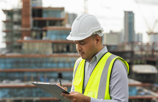 Inspector engineer man or architect with clipboard with white safety helmet in city construction site. Checking with checklist on rooftop building construction at capital. stock photo