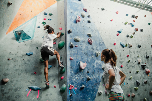 Two people are enjoying an intense climbing session at an indoor rock climbing centre. The man is climbing a wall and his friend is watching him with her hands on her hips.