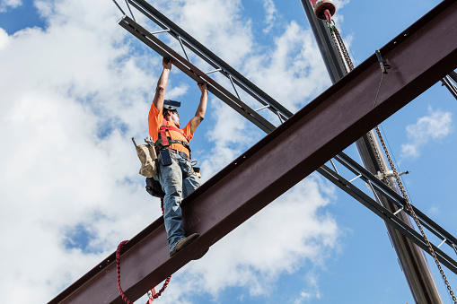 An Hispanic steel worker working high up on a girder. He is standing on the girder, wearing a safety harness, reaching up for a roof joist or truss which is being lifted into place by a crane. He has a welding helmet on his head.