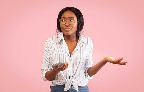 Irritated Girl Holding Smartphone Shrugging Shoulders On Pink Background Irritated African American Girl Holding Smartphone And Shrugging Shoulders Looking Aside Standing On Pink Background. Studio Shot irritation stock pictures, royalty-free photos & images