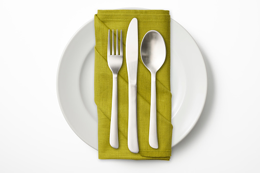 Overhead shot of white dinner plate and cutlery with green napkin, isolated on white background.