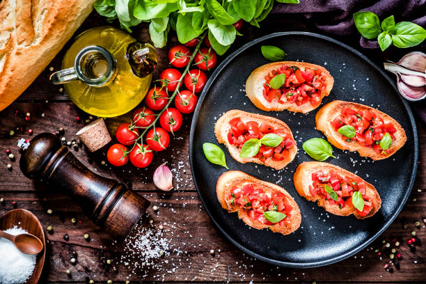 Italian bruschetta Top view of a homemade Italian bruschetta made with cherry tomatoes, basil, olive oil, garlic and salt disposed on a black plate surrounded by the ingredients for preparing the bruschetta on a dark brown wooden table. Low key DSLR photo taken with Canon EOS 6D Mark II and Canon EF 24-105 mm f/4L antipasto stock pictures, royalty-free photos & images