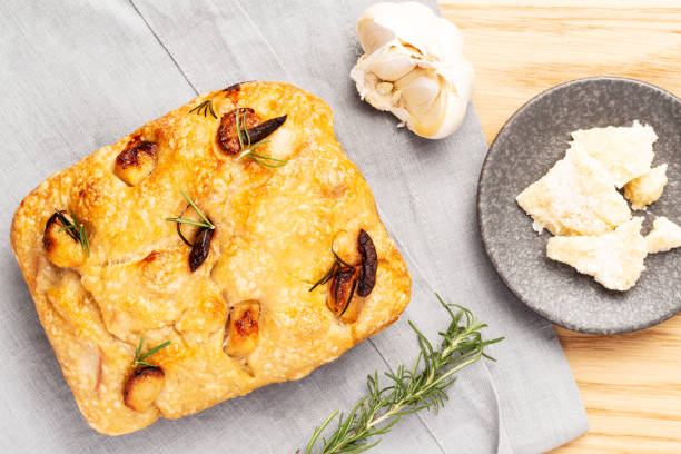 Italian focaccia with confit garlic cloves and rosemary, alongside a golden dessert tong, garlic and parmesan cheese on a grey napkin on a wooden table. Top view. stock photo