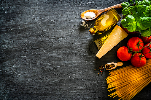 Italian food background: overhead view of a black table with uncooked spaghetti and ingredients for cooking a traditional Italian pasta arranged at the right of a black background making a frame and leaving useful copy space. The composition includes basil. fresh ripe tomatoes, olive oil, garlic, pepper, salt and Parmesan cheese. High resolution 42Mp studio digital capture taken with Sony A7rII and Sony FE 90mm f2.8 macro G OSS lens