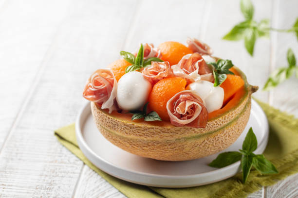 Italian summer salad. Prosciutto, mozzarella cheese and melon balls in melon basket, decorated with basil leaves. White background, copy space. Selective focus. stock photo